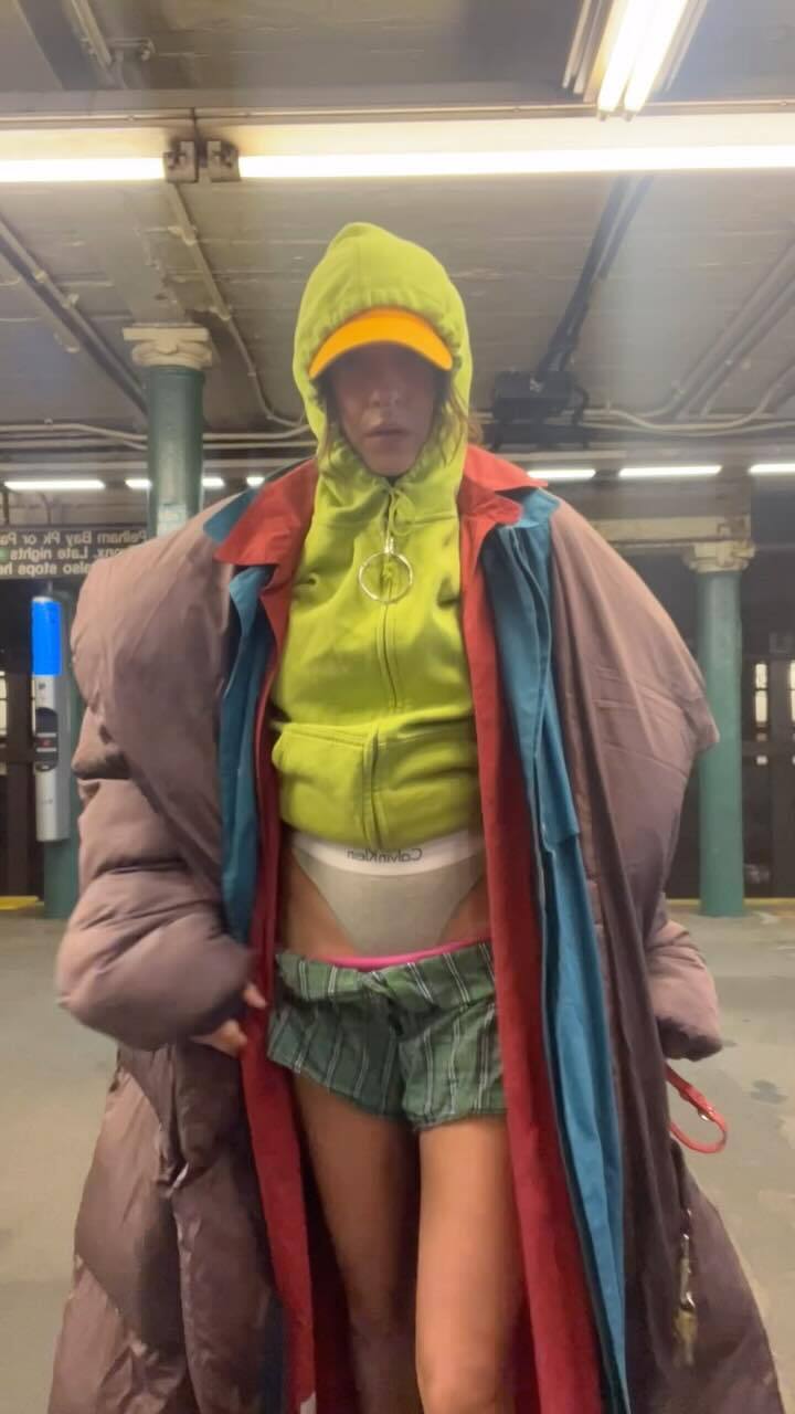 Person in layered clothing, including a green hoodie, orange cap, and oversized coat, stands in a subway station.