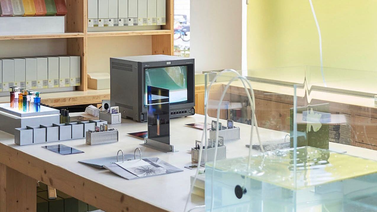 The Broken Arm Paris, A modern laboratory setup with various equipment, including a monitor, binders on shelves, test tubes with colorful liquids, and a glass tank.