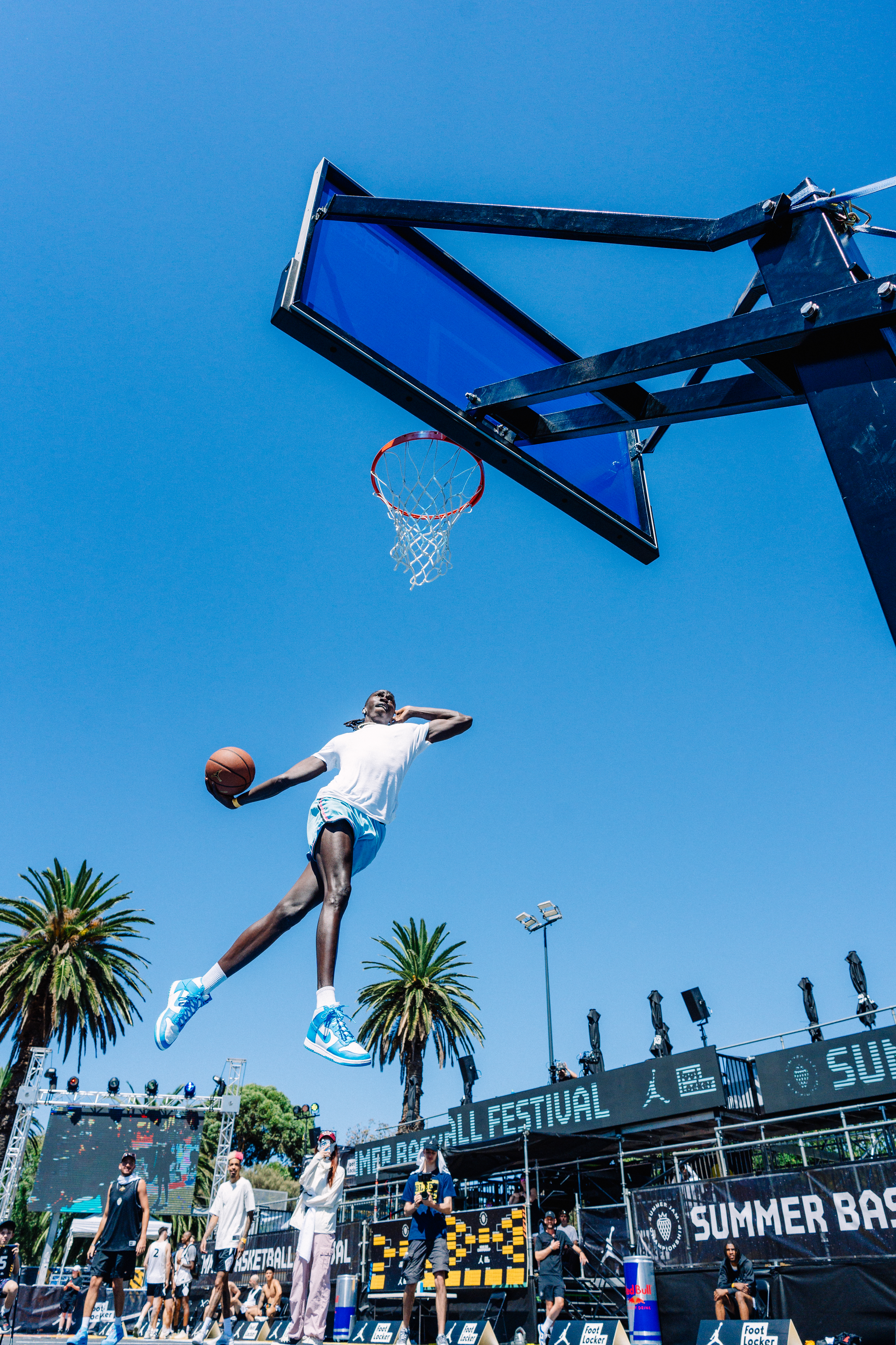 A basketball player in mid-air, performing a slam dunk at an outdoor court with a clear blue sky and palm trees in the background.