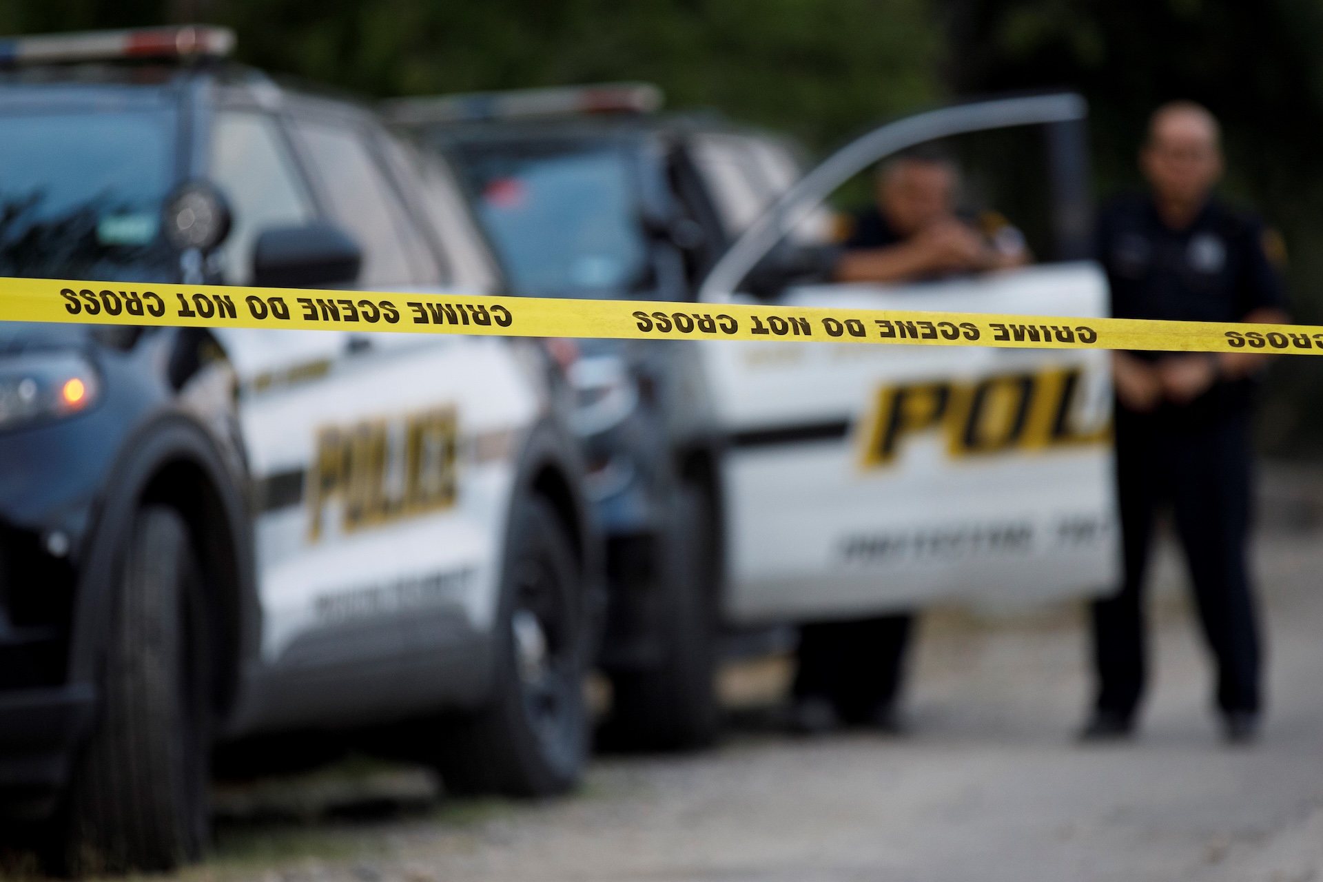 Police cars behind yellow "Crime Scene Do Not Cross" tape, with two officers standing in the background.