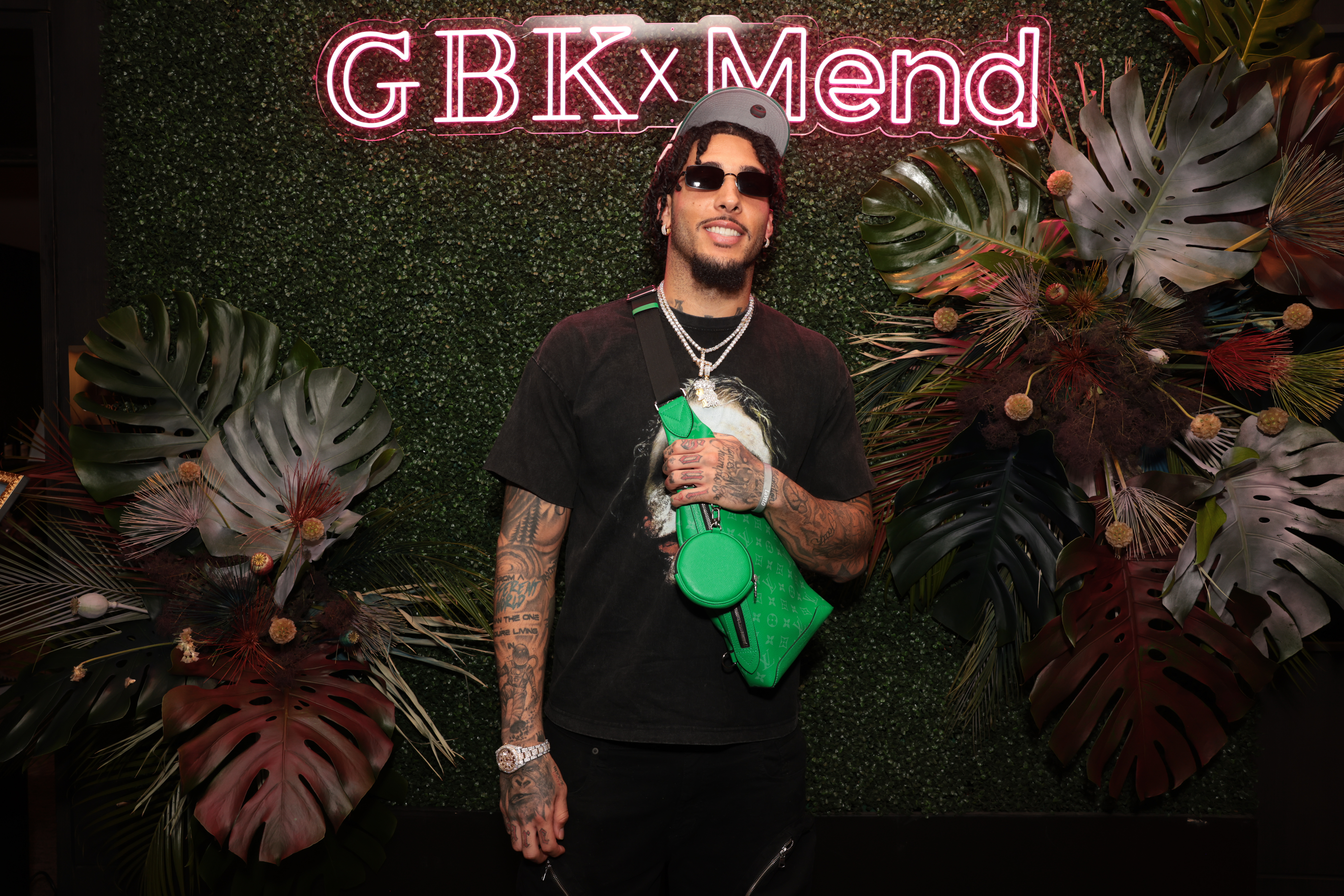 LiAngelo Ball with tattoos and sunglasses stands in front of a "GBK x Mend" sign, wearing a black shirt and a green crossbody bag, surrounded by tropical leaves.