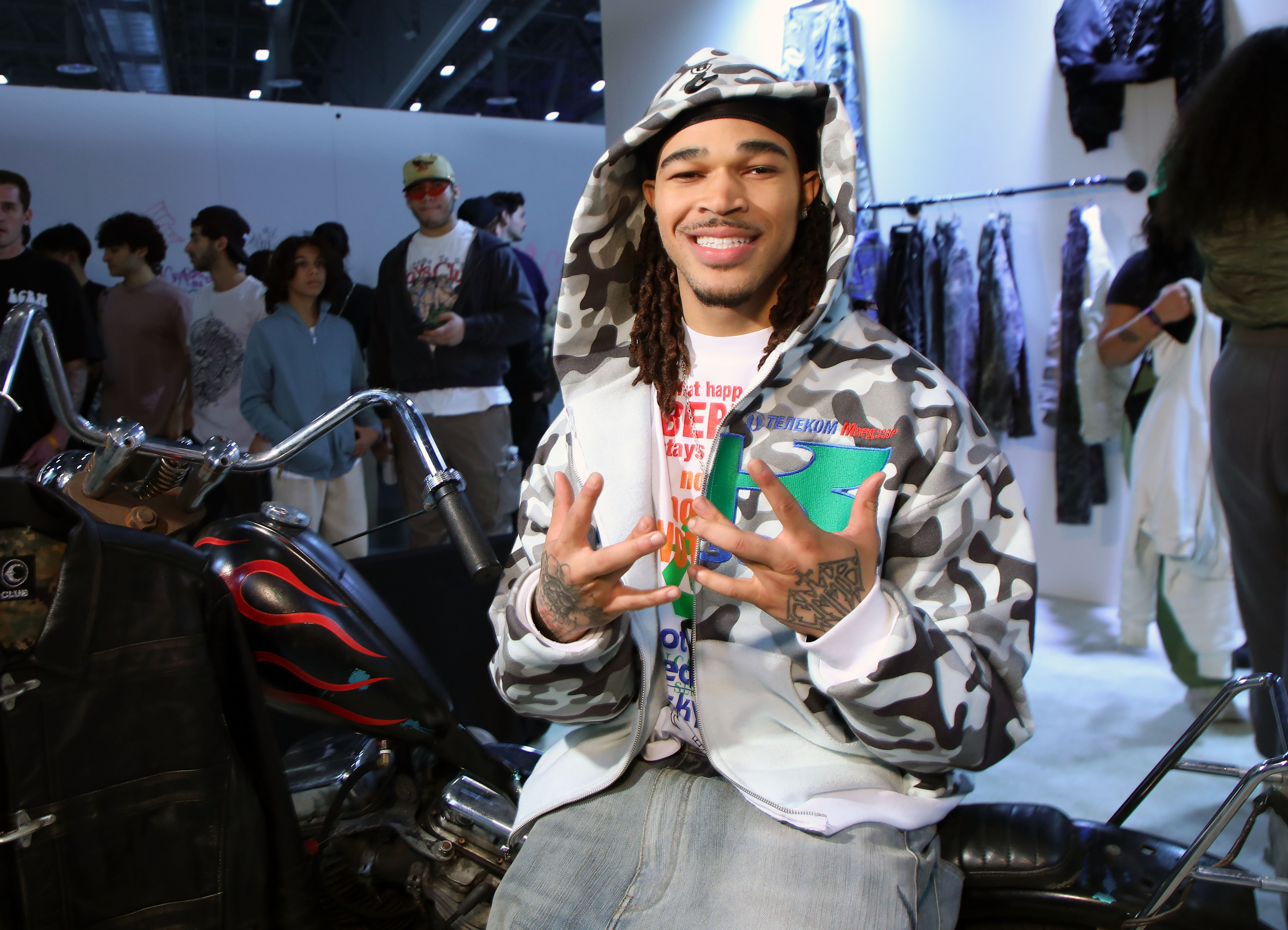 A person in a camo hoodie sits on a motorcycle at a fashion event, smiling and holding up hand signs. People and clothing racks are in the background.