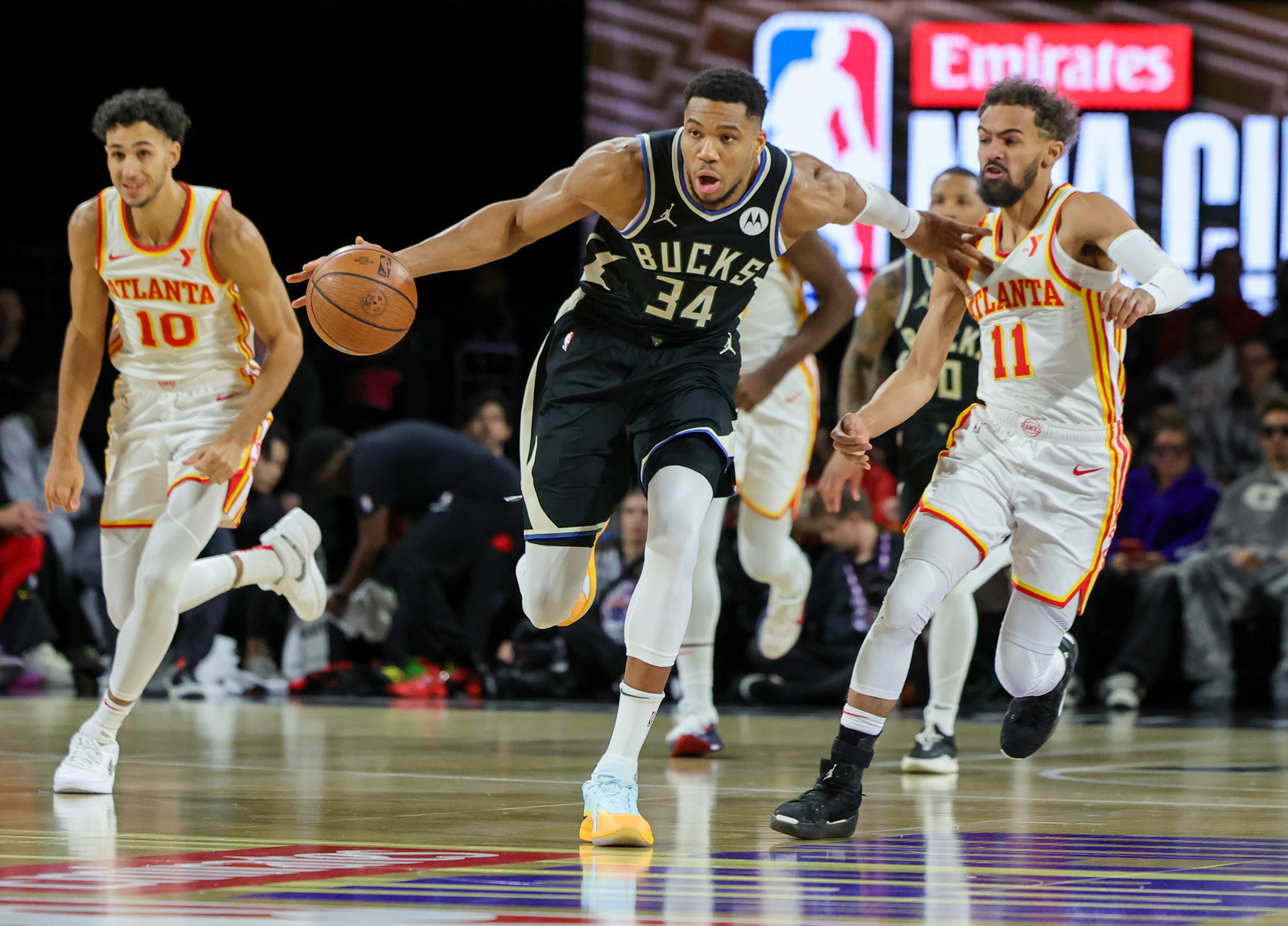 Giannis Antetokounmpo dribbles the basketball, flanked by Atlanta Hawks players Trae Young and another teammate during an NBA game.