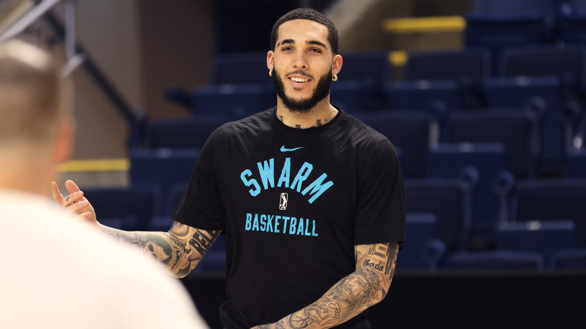 LiAngelo with tattoos is wearing a "Swarm Basketball" shirt, smiling on the court.